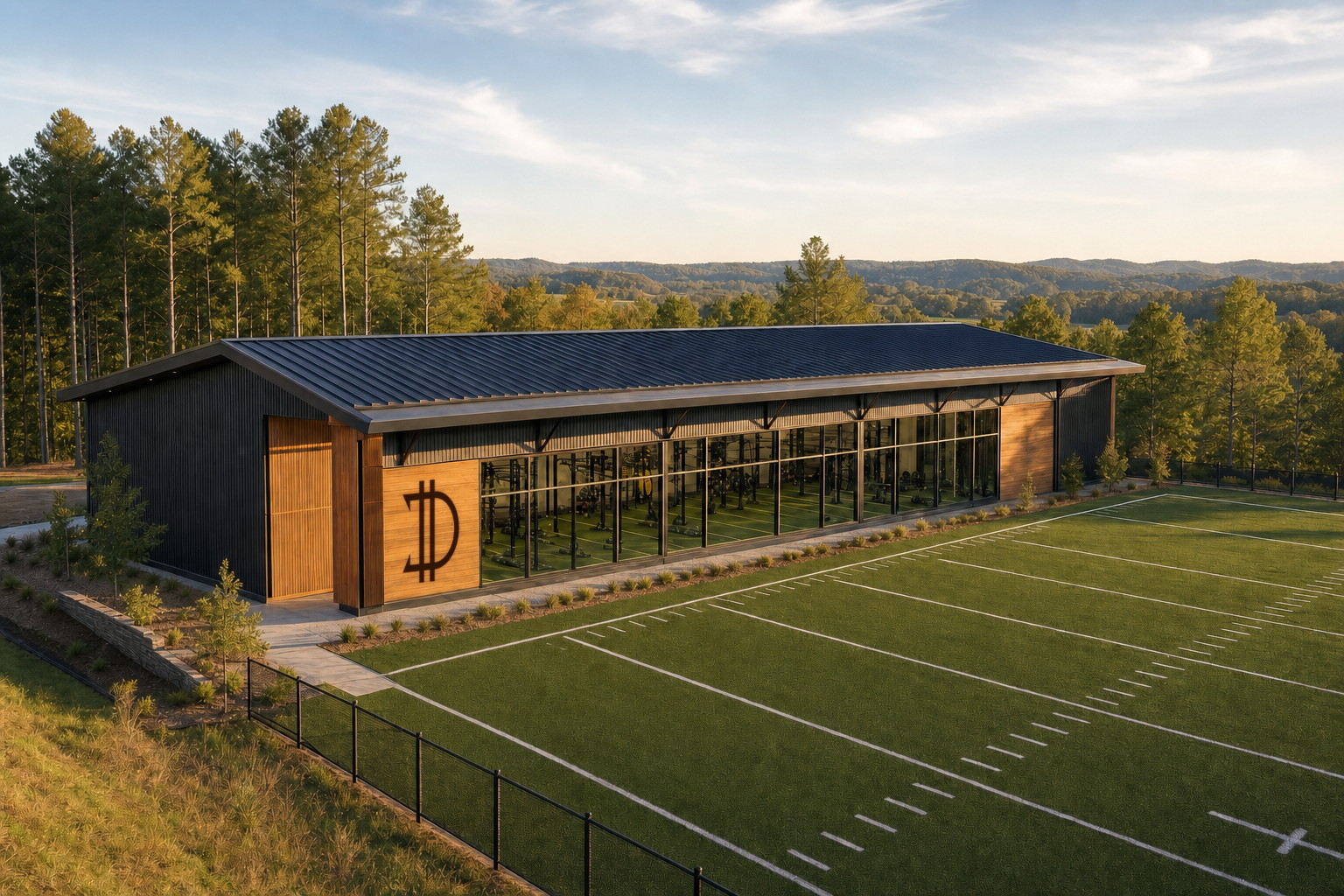A concept illustration of a low modern training building in charcoal metal and warm wood with a glass training bay, fronted by a full outdoor turf field with white yard lines, pines and rolling hills behind it.